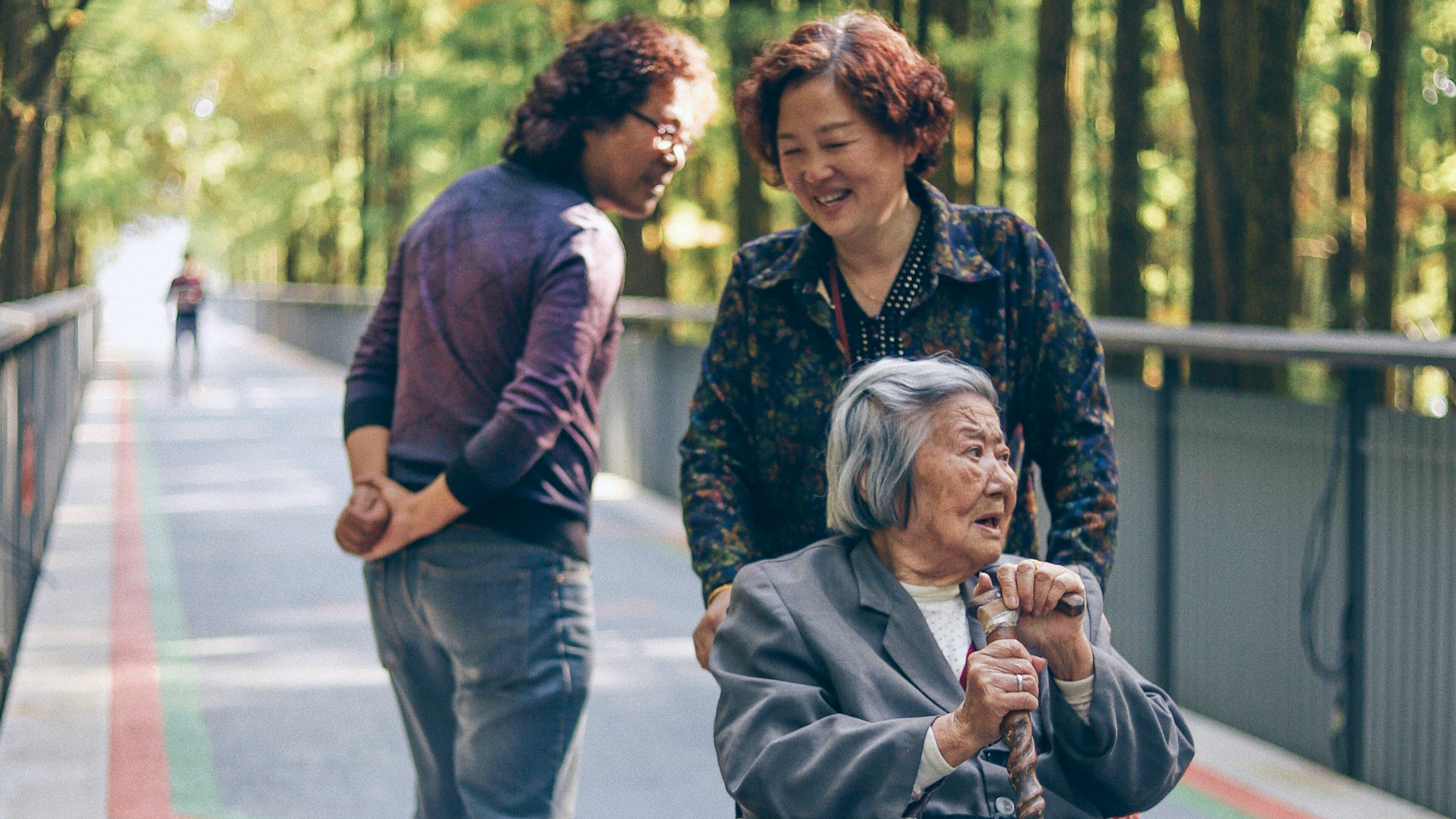 An Asian woman stops to talk to a man while pushing an older Asian woman in a wheel chair along a paved wooded path.