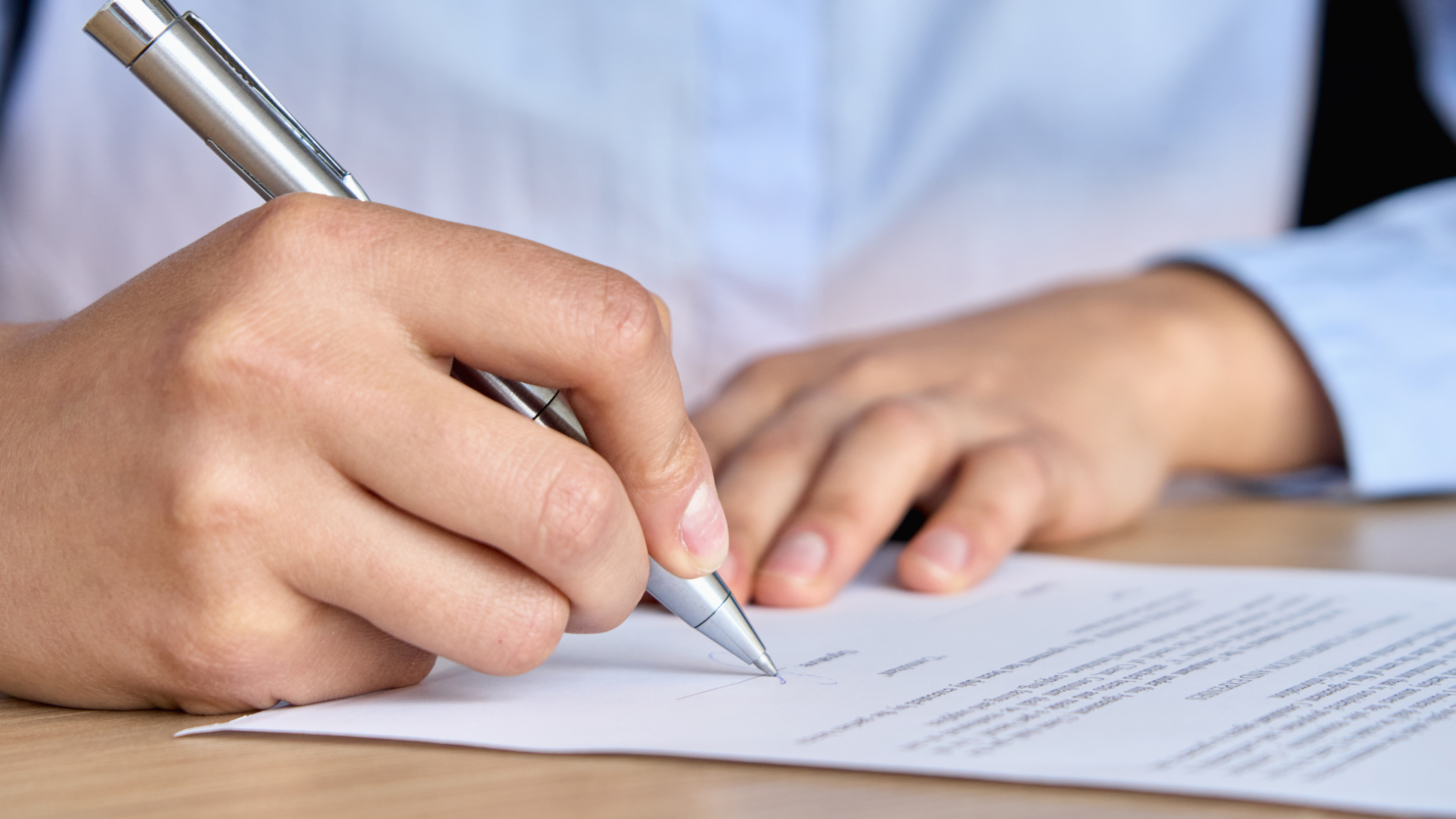 A person in a blue shirt is writing with a silver pen on a piece of paper.