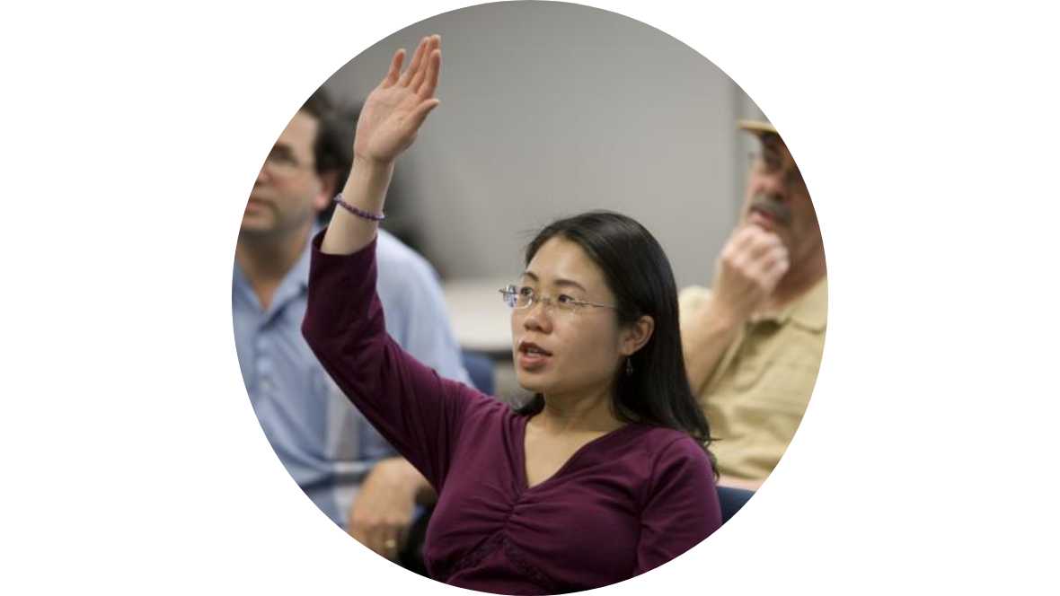 One woman in a dark red top with her raised hand while 2 men are sitting behind her.