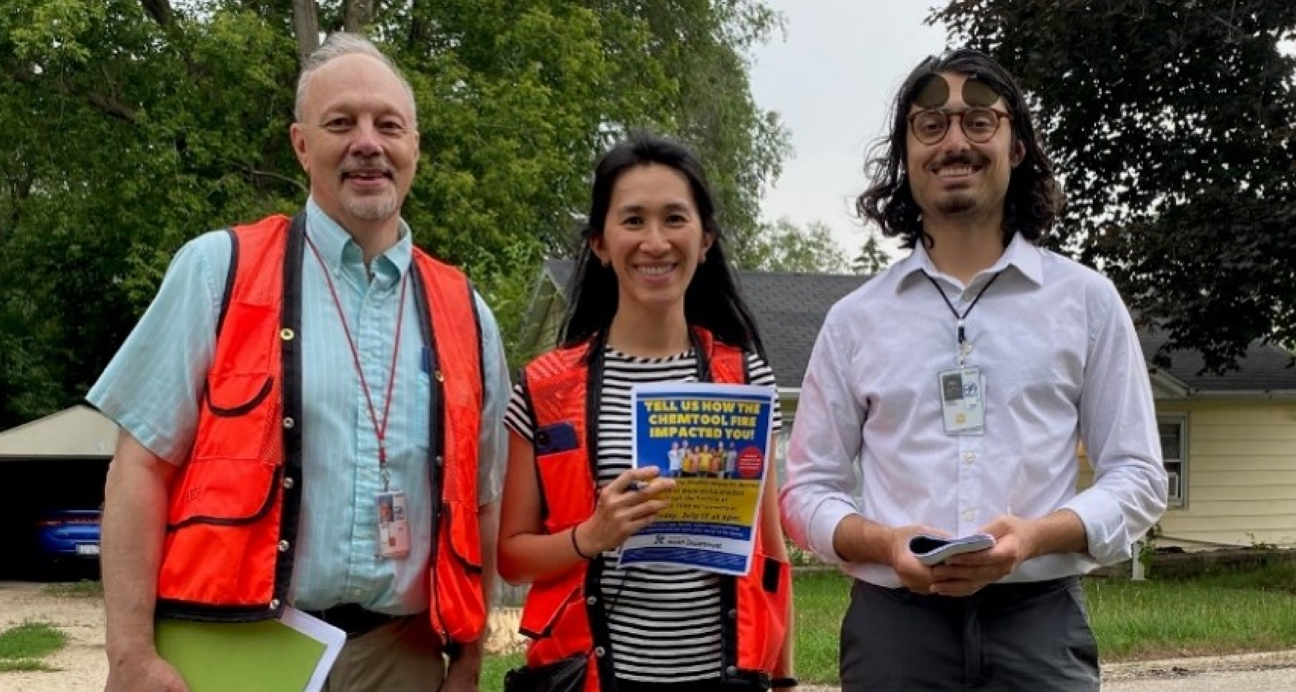 Three people, standing and smiling together, two with orange vests on holding papers.