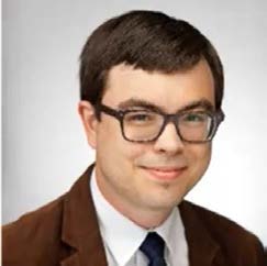 A man with short brown hair, square-framed glasses in a brown suit jacket and white-collar shirt with a black tie.