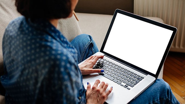 A woman with a blue and white shirt and blue jeans sitting on a couch looking at a blank laptop screen.