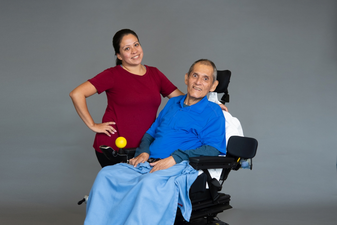 A woman in a dark red shirt standing next to man in a blue shirt with ALS in wheelchair.