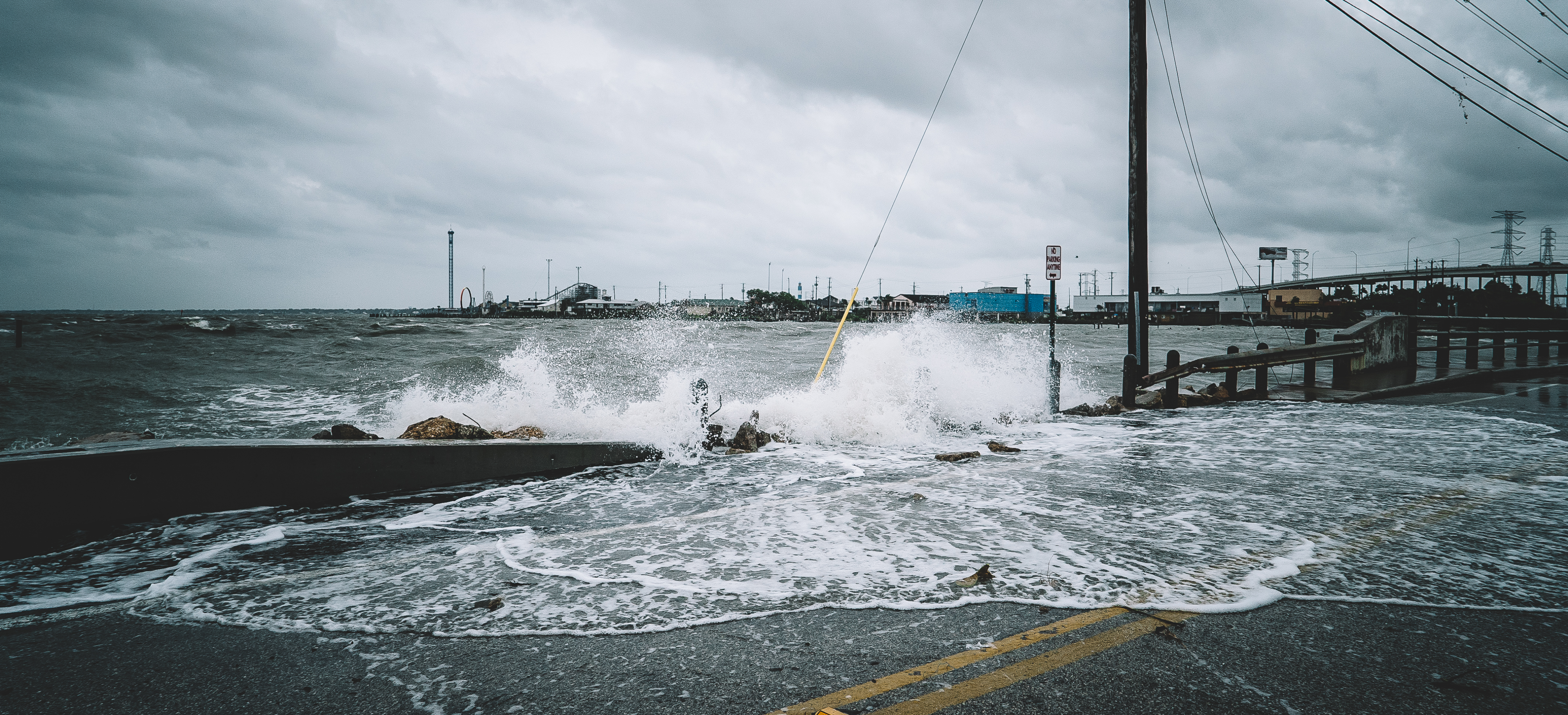 Water flooding into the street over cement and a fence with dark clouds in the background.