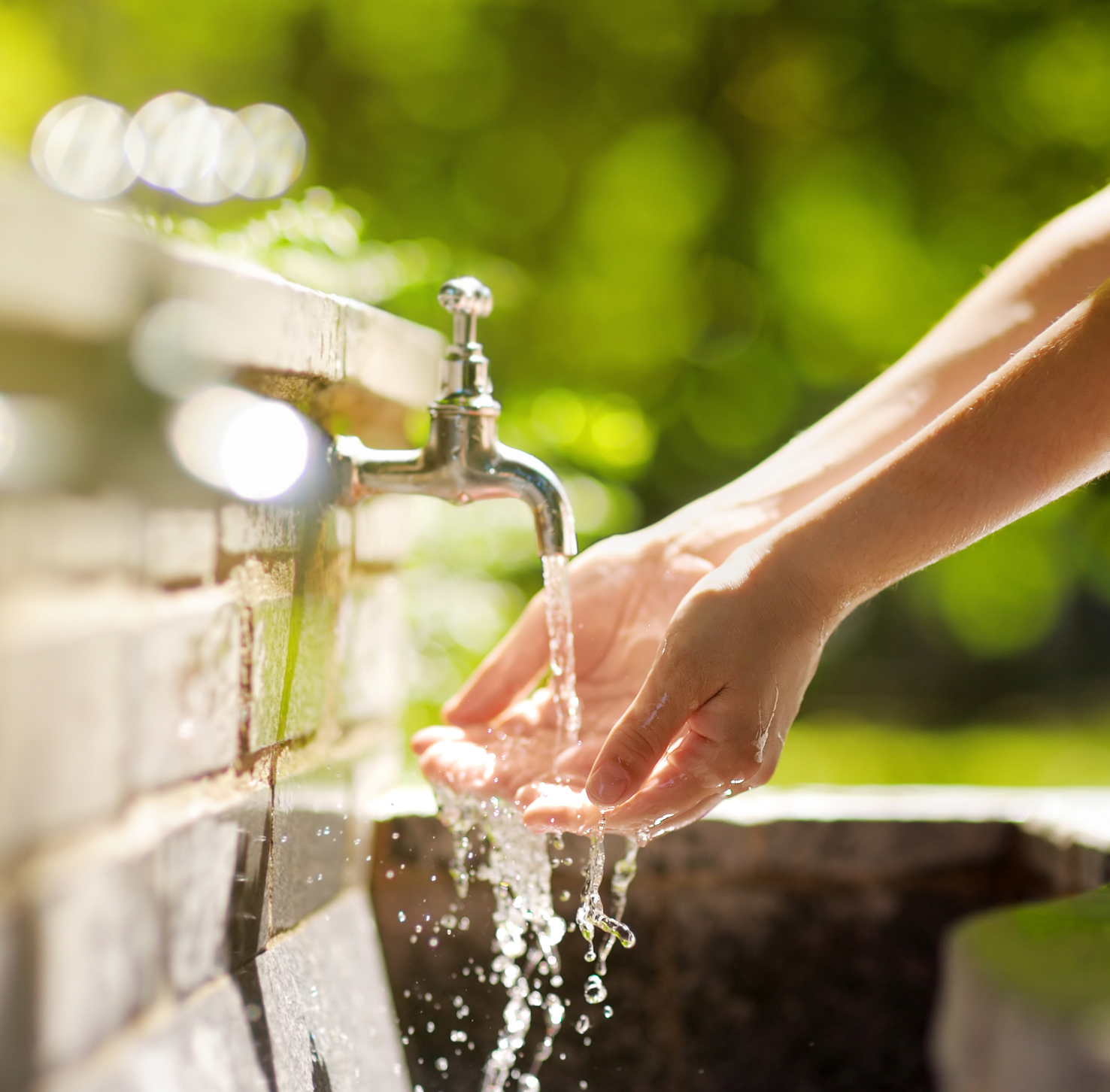 Two hands washing in an outdoor sink made of brick with greenery in the background.