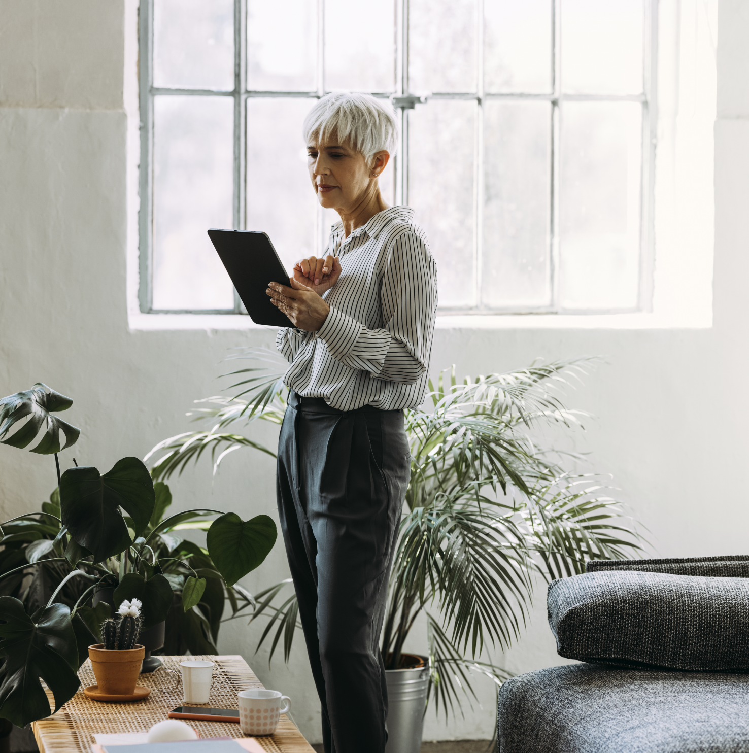 A woman with short white hair reading a clipboard with a window and plant in the background.