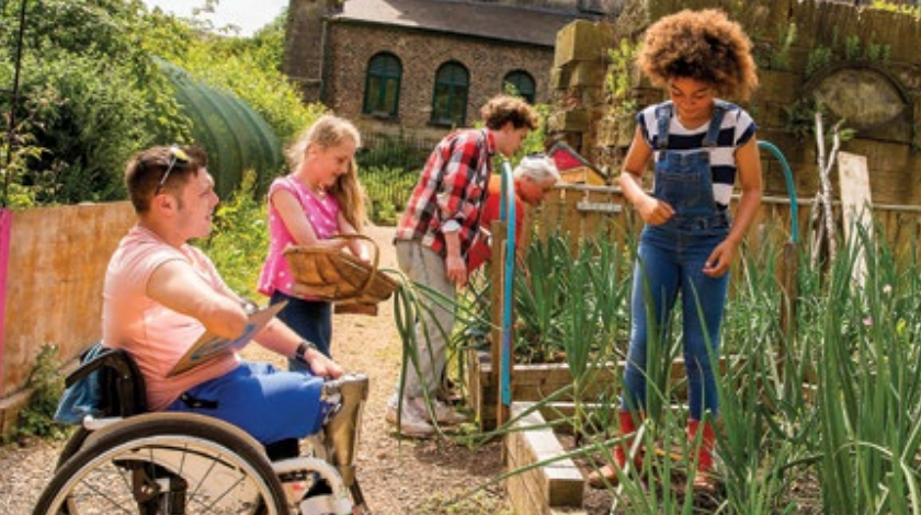 A man in a wheelchair, a girl in overalls and boots, and others outside in a garden.