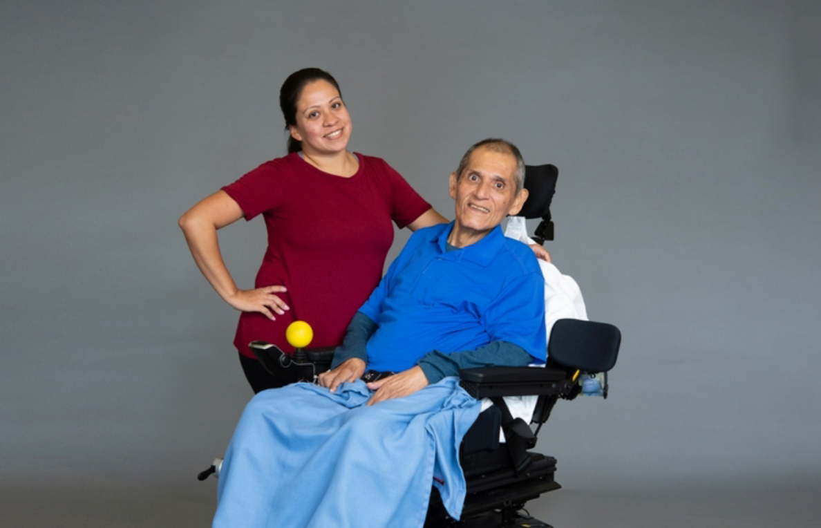 A woman in a dark red shirt standing next to a man in a blue shirt and blue jeans in a wheelchair.