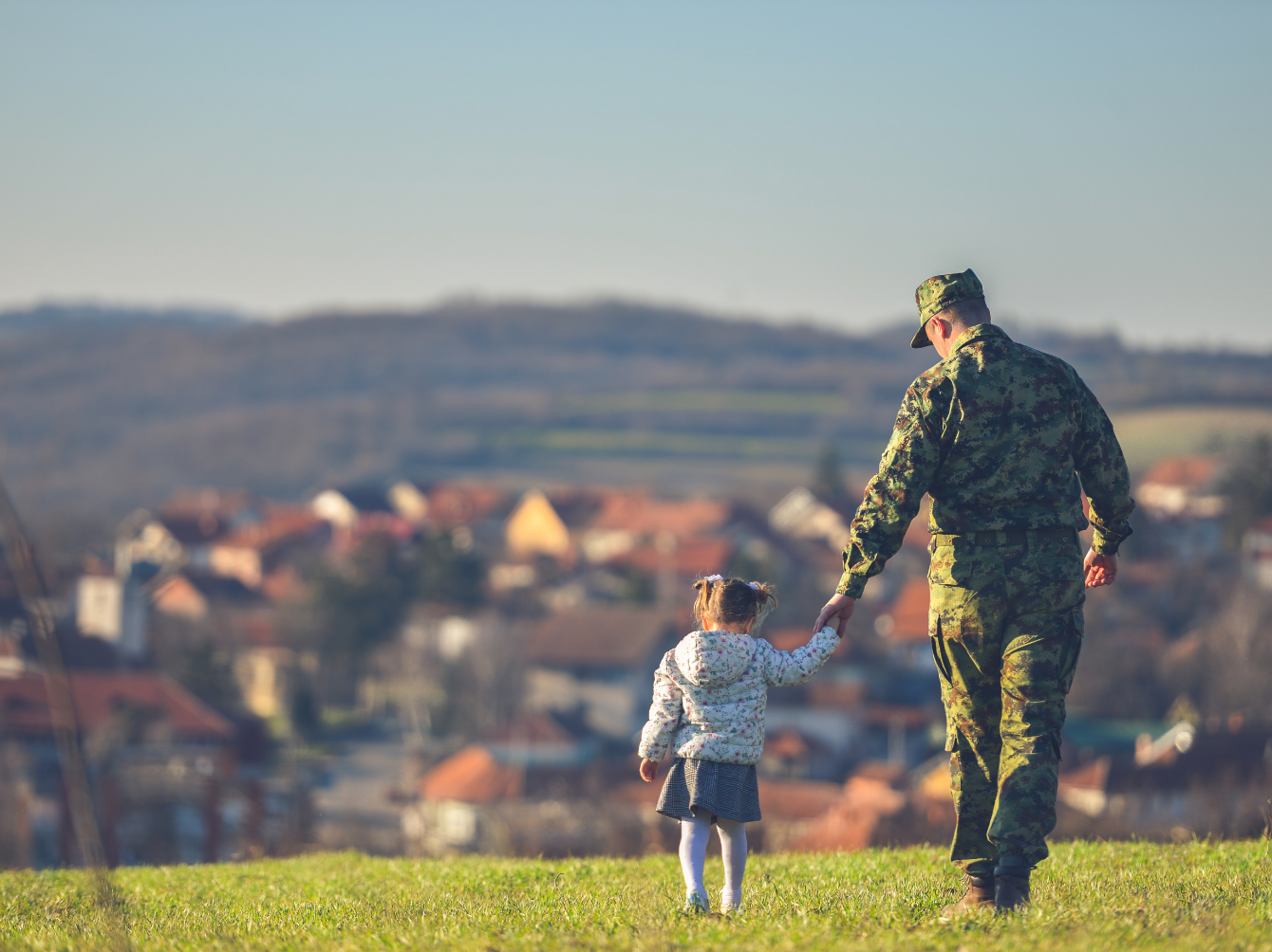 A man wearing a military uniform is walking and holding the hand of a little girl in a field with houses in the background.