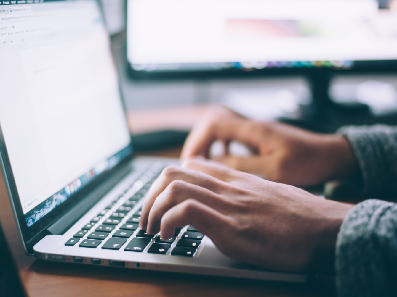 Two hands typing on a laptop with a computer monitor in the background.