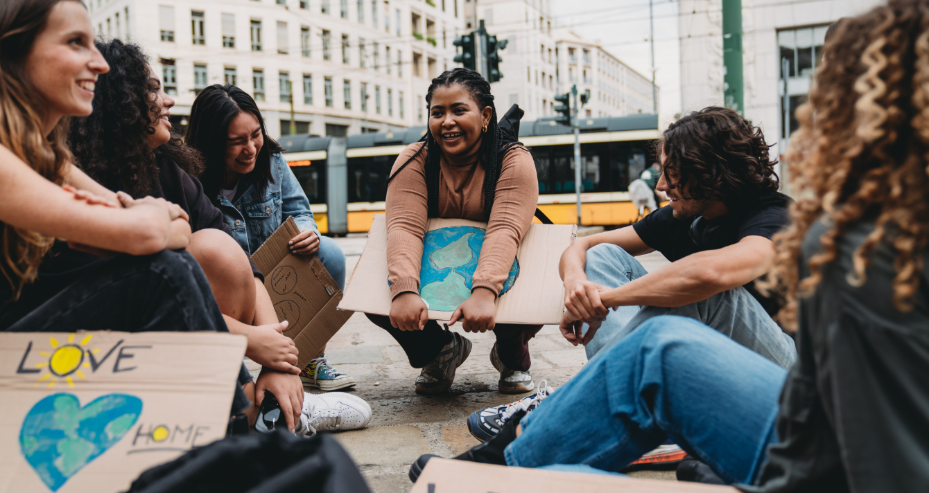 Six people sitting in a circle holding signs showing the world with buildings behind them.