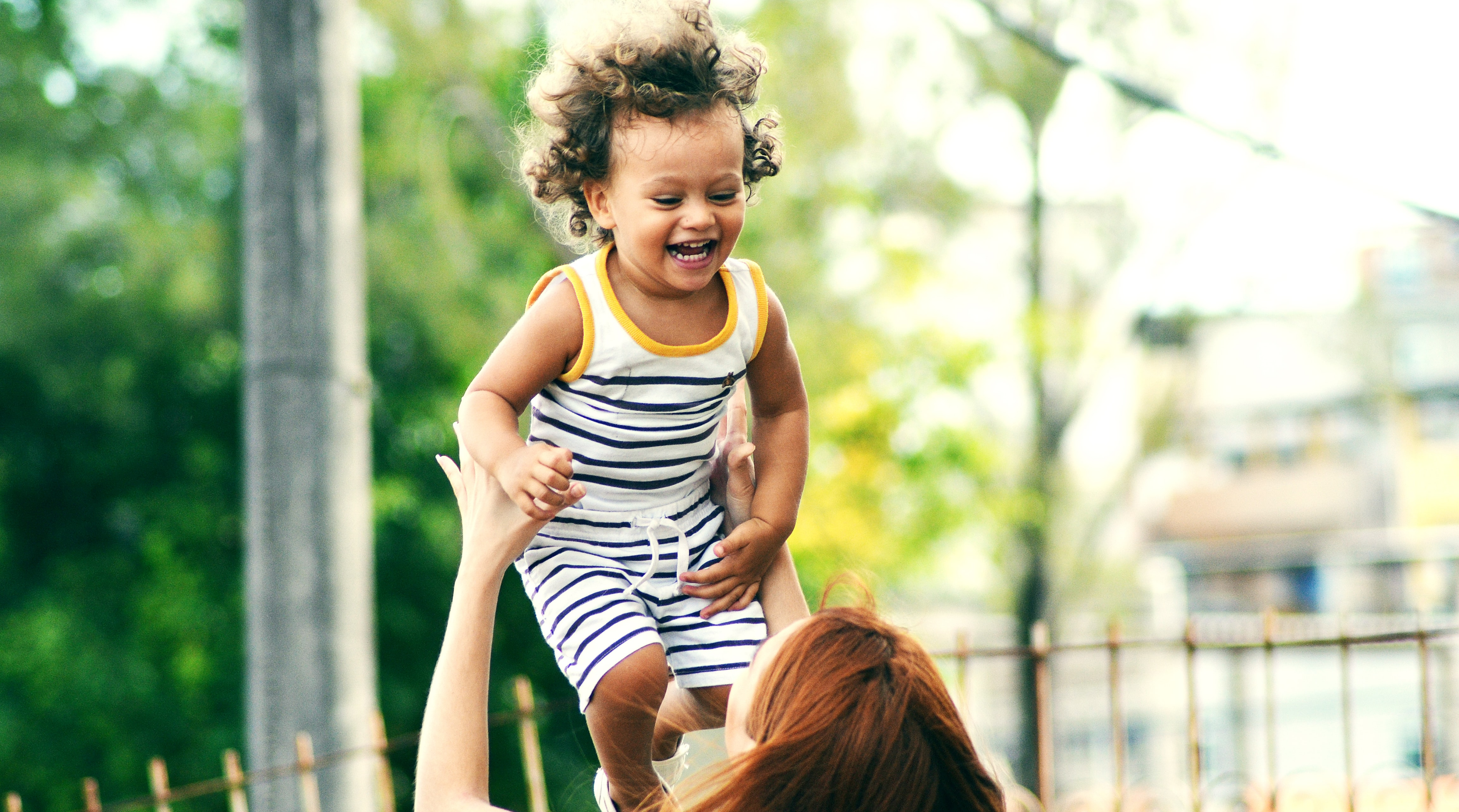 A woman with red hair is throwing a laughing child wearing a striped shirt and shorts in the air.