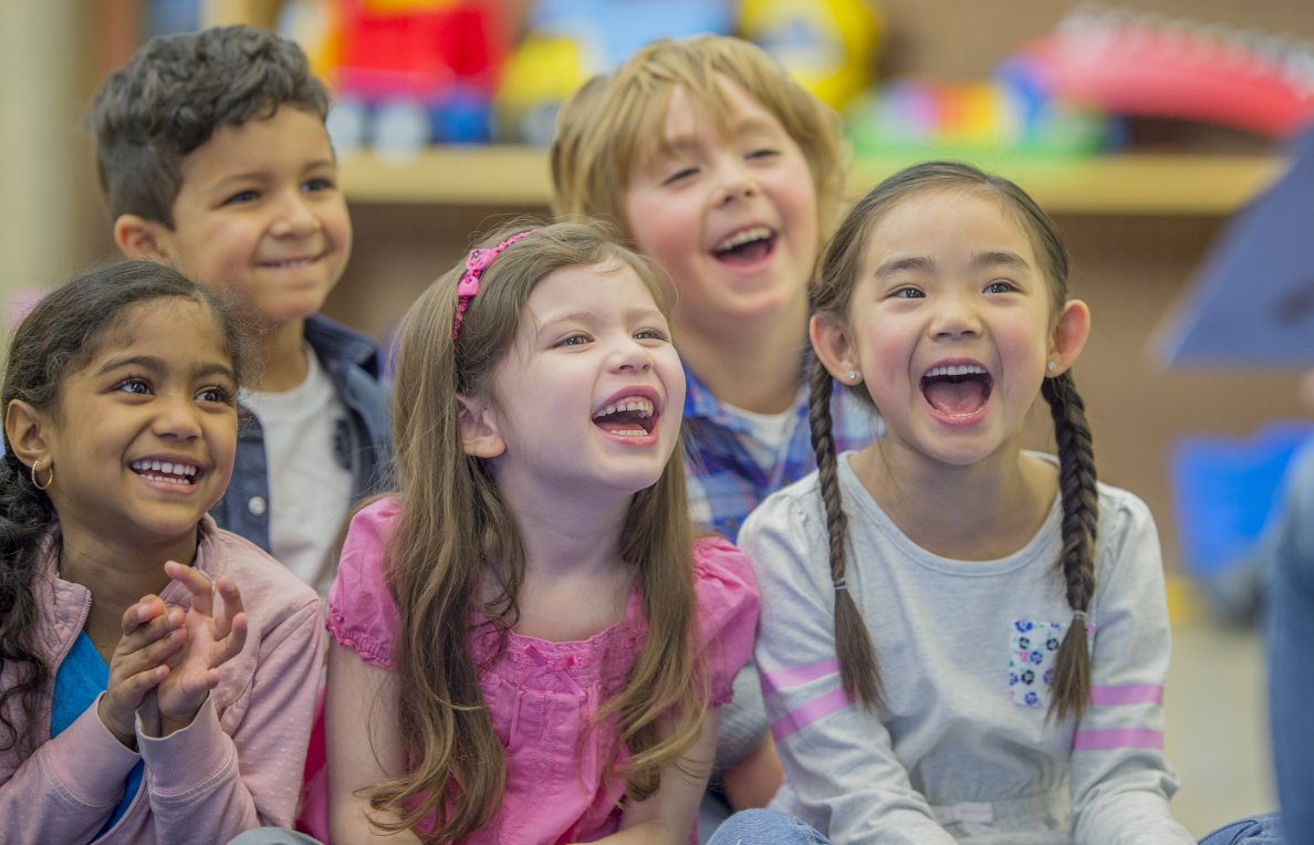 A group of 5 children sitting down smiling and laughing with colorful books in the background.