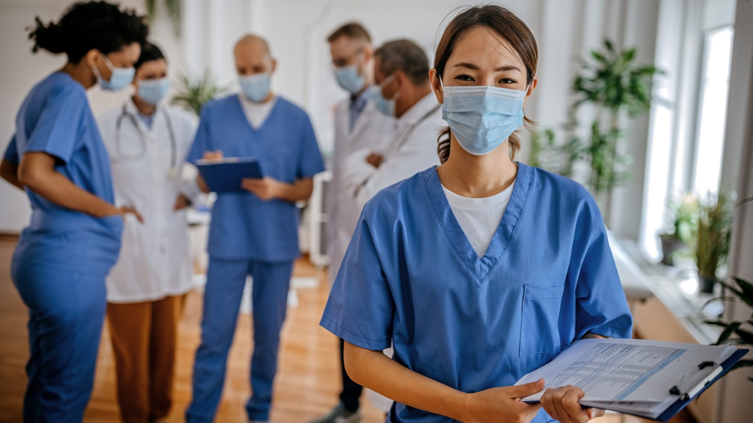 A diverse team of six healthcare providers wearing scrubs, medical coats, and masks, in a discussion.