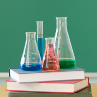 Four flasks filled with different colored chemicals in four flasks on books against a green background.