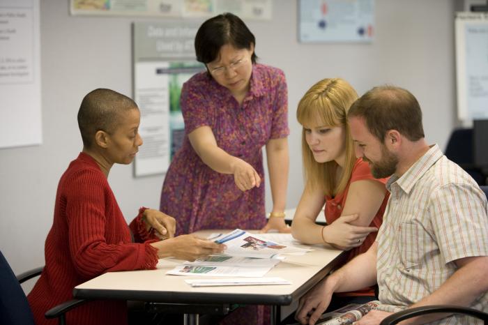 Four people are gathered around a table. One person is pointing at a document on the table that everyone is looking at.