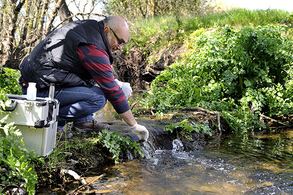 Man, in navy vest, red and navy stiped shirt, and jeans, who is an epidemiologist, collects water samples by stream.