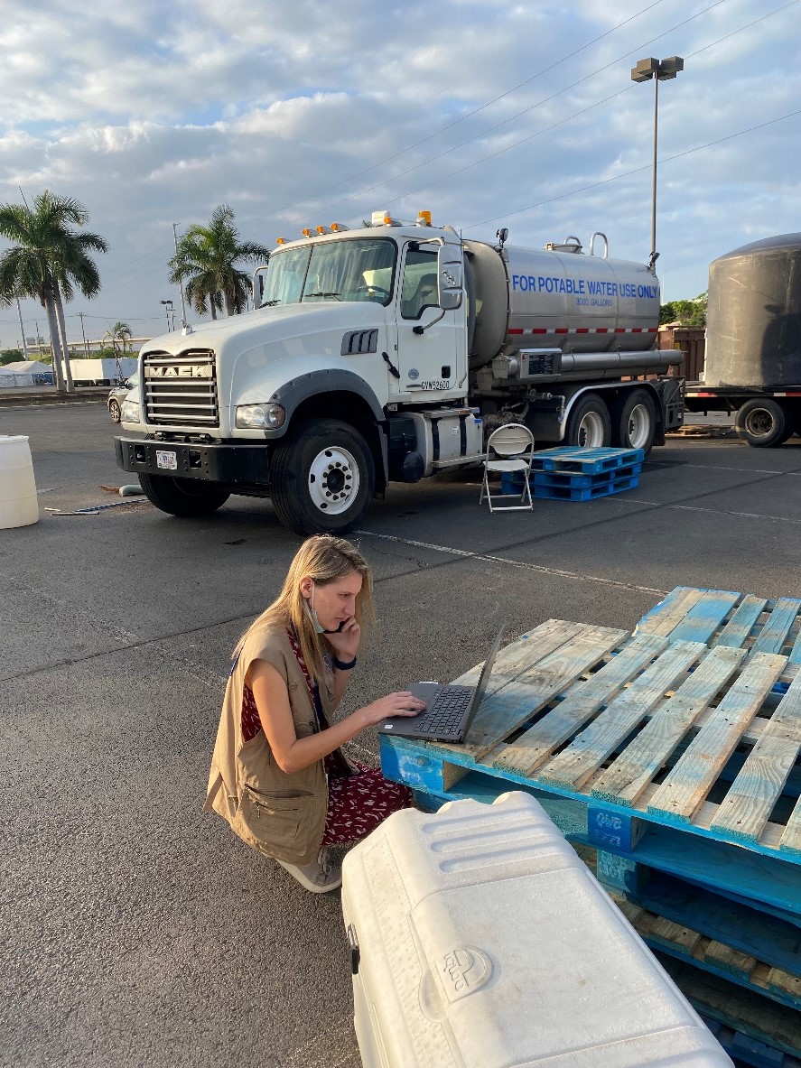 ACE team member works on laptop on stacked pallets next to portable water truck.